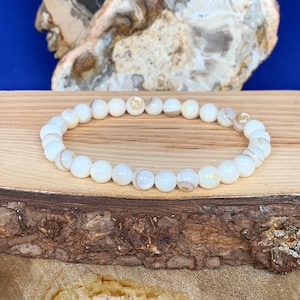 Op de afbeelding: Een armband van witte schelp kralen met bruine en witte patronen op elke kraal. De armband ligt op een houten oppervlak.