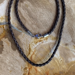 Peut inclure: Un collier en cuir tressé marron foncé avec un fermoir argenté. Le collier est présenté sur une surface texturée brun clair. Le fermoir est de type mousqueton.