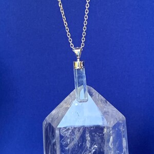 May include: A silver-toned chain necklace featuring a clear, cylindrical crystal pendant with a metallic cap. The pendant is positioned directly in front of a large, transparent, multi-faceted quartz crystal point with visible internal fractures.