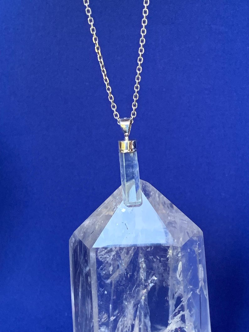 May include: A silver-toned chain necklace featuring a clear, cylindrical crystal pendant with a metallic cap. The pendant is positioned directly in front of a large, transparent, multi-faceted quartz crystal point with visible internal fractures.
