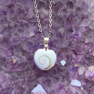 Op de afbeelding: Een zilveren ketting met een witte hartvormige hanger met een spiraalvormig schelpmotief. De ketting wordt getoond op een bed van paarse amethistkristallen.