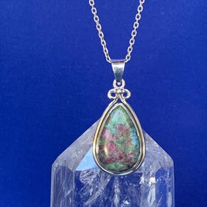 May include: A silver pendant necklace with a teardrop-shaped stone displaying green, red, and blue colours. The stone is set in a silver bezel and suspended from a delicate silver chain. The background is a solid blue.