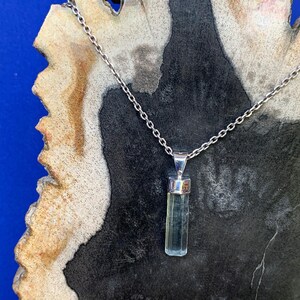 May include: A silver chain necklace featuring a vertical, pale green, translucent, rectangular crystal pendant held by a polished silver cap. The pendant hangs from a delicate metal link chain, showcasing a clear, faceted, elongated gemstone design.