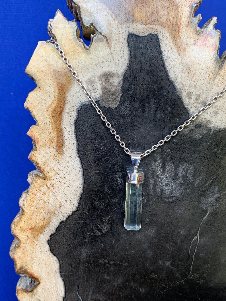 May include: A silver chain necklace featuring a vertical, pale green, translucent, rectangular crystal pendant held by a polished silver cap. The pendant hangs from a delicate metal link chain, showcasing a clear, faceted, elongated gemstone design.