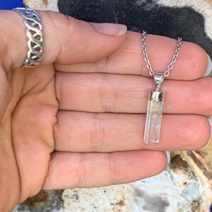 May include: A silver chain necklace featuring a clear, rectangular, faceted crystal pendant with a silver cap, accompanied by a silver ring with an open, woven, or lattice-style band design, both showcasing a polished metallic finish.