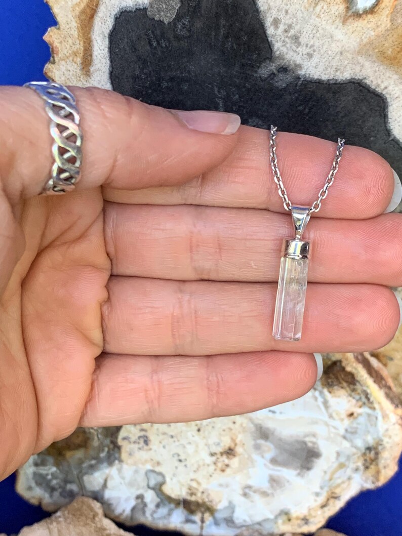 May include: A silver chain necklace featuring a clear, rectangular, faceted crystal pendant with a silver cap, accompanied by a silver ring with an open, woven, or lattice-style band design, both showcasing a polished metallic finish.