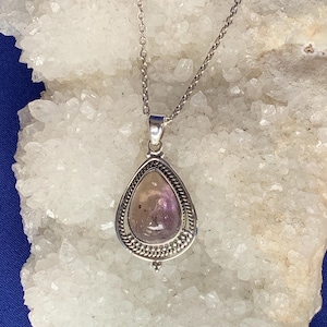 May include: A silver necklace with a teardrop-shaped pendant. The pendant has a light purple gemstone set in a detailed silver frame. The chain is delicate and the pendant hangs gracefully.