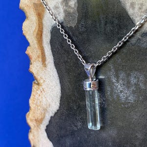 May include: A silver-toned metal chain necklace featuring a vertical, translucent, pale blue cylindrical crystal pendant held by a polished silver-toned metal cap and bail. The pendant hangs straight down from the delicate link chain.