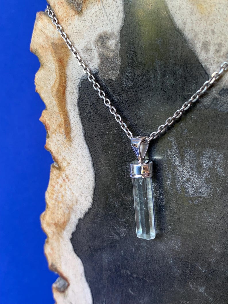 May include: A silver-toned metal chain necklace featuring a vertical, translucent, pale blue cylindrical crystal pendant held by a polished silver-toned metal cap and bail. The pendant hangs straight down from the delicate link chain.