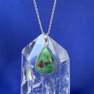 May include: A silver pendant necklace with a triangular gemstone. The gemstone has a green base with red inclusions. The pendant hangs from a delicate silver chain. The necklace is displayed on a clear crystal against a blue backdrop.