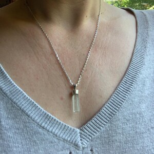 May include: A silver-toned chain necklace featuring a vertical, rectangular, pale green translucent pendant. The pendant is capped with a metallic silver-toned bail that connects it to the delicate link chain, resting against a light gray knit fabric.