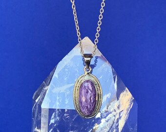 Peut inclure: Un collier en chaîne argent avec un pendentif ovale violet et blanc. Le pendentif est serti dans un cadre argenté avec une bordure décorative. Le collier est présenté sur un cristal transparent.