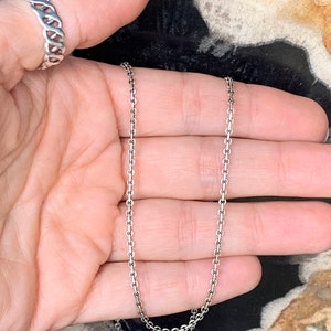May include: A silver chain necklace with a delicate, fine link design. The chain is shown lying on a hand, with the ends of the chain hanging down.