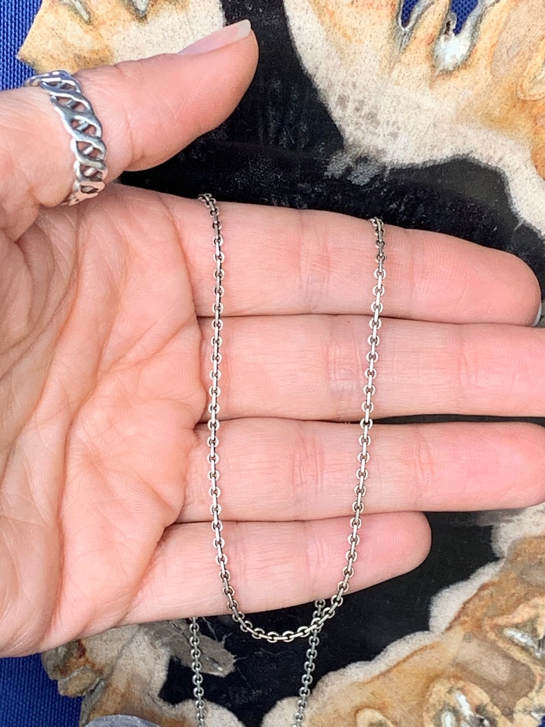May include: A silver chain necklace with a delicate, fine link design. The chain is shown lying on a hand, with the ends of the chain hanging down.