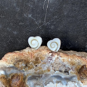 Op de afbeelding: Twee hartvormige oorbellen met een wit spiraalontwerp, gepresenteerd op een getextureerde, veelkleurige steen. De donkere achtergrond accentueert het unieke ontwerp van de oorbellen.