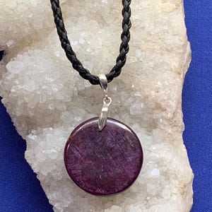 May include: A circular, deep purple-red gemstone pendant with a smooth, polished surface, suspended from a black braided cord by a silver-toned metal bail. The pendant features natural internal inclusions and a rich, dark color profile.