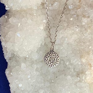 Op de afbeelding: Een zilverkleurige metalen ketting met een ronde hanger met een ingewikkeld opengewerkt ontwerp en een centraal Om-symbool. De hanger hangt aan een fijne schakelketting en toont een gedetailleerd, gestructureerd oppervlak op de hanger.