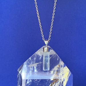 May include: A silver-toned metal chain necklace featuring a vertical, rectangular, pale blue translucent stone pendant with a silver-toned metal cap. The pendant is suspended from a delicate link chain, showcasing a clear, faceted crystal structure.
