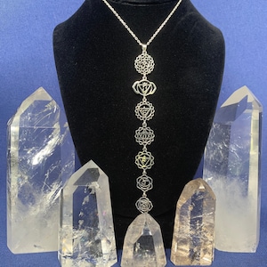 Peut inclure: Un collier en argent avec sept symboles de chakras suspendus. Le collier est présenté sur un support en velours noir avec des cristaux de quartz transparents de chaque côté.