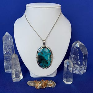 Peut inclure: Un collier pendentif en argent avec une pierre ovale turquoise, présenté sur un buste blanc. Des cristaux de quartz clair entourent le collier. La pierre du pendentif présente des nuances de bleu et de vert.