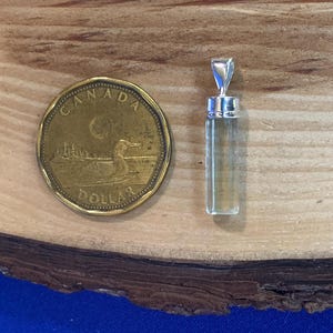 May include: A clear, cylindrical crystal pendant with a silver-toned metal cap and bail, positioned next to a gold-colored Canadian one-dollar coin for scale. The pendant features a smooth, faceted surface and a transparent, light-toned appearance.