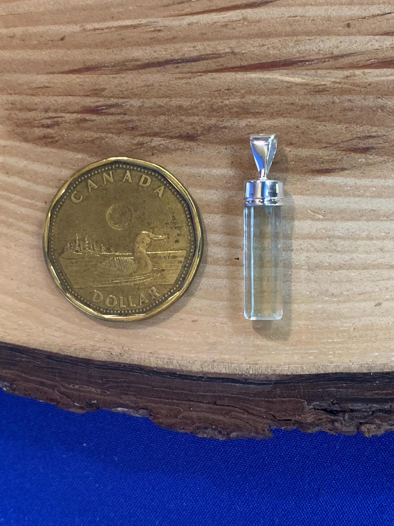 May include: A clear, cylindrical crystal pendant with a silver-toned metal cap and bail, positioned next to a gold-colored Canadian one-dollar coin for scale. The pendant features a smooth, faceted surface and a transparent, light-toned appearance.
