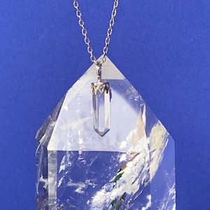May include: A clear quartz crystal pendant on a silver chain, displayed on a clear quartz crystal point.