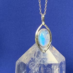 May include: A silver necklace featuring an oval moonstone pendant with a blue iridescent sheen, set within a pointed, eye-shaped metal frame. The pendant hangs from a delicate silver chain, positioned above a clear, faceted quartz crystal point.