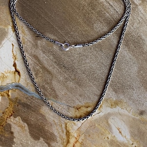 Peut inclure: Un collier argenté avec un motif de chaîne tressée. Le collier est présenté sur une surface brun clair. La chaîne est reliée par un petit fermoir. Il s'agit d'un bijou.