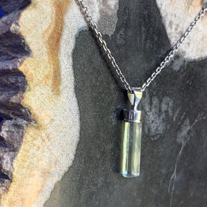 May include: A pendant necklace featuring a long, cylindrical, pale yellow-green crystal suspended from a silver-toned metal cap. The crystal is attached to a delicate silver-toned metal chain that extends upwards from the top of the pendant.