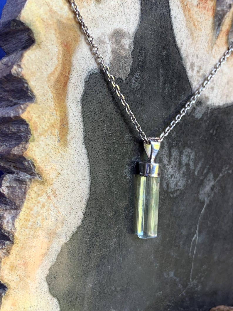 May include: A pendant necklace featuring a long, cylindrical, pale yellow-green crystal suspended from a silver-toned metal cap. The crystal is attached to a delicate silver-toned metal chain that extends upwards from the top of the pendant.