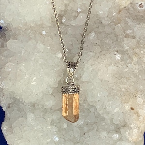 May include: A silver-toned metal chain necklace featuring a vertical, translucent, light orange crystal pendant. The crystal is held by a decorative, textured silver-toned metal cap with a small loop connecting it to the delicate link chain.