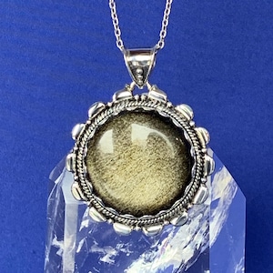 May include: A silver necklace featuring a circular, gold-toned pendant. The pendant has a textured surface and is set in a decorative silver frame. The necklace is displayed on a clear, crystal-like object against a blue backdrop.