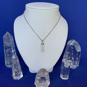 May include: A pale blue, cylindrical crystal pendant hangs from a thin, silver-toned metal chain. The pendant is capped with a small silver-toned metal bail. The necklace is displayed on a white, neck-shaped stand against a solid blue background.