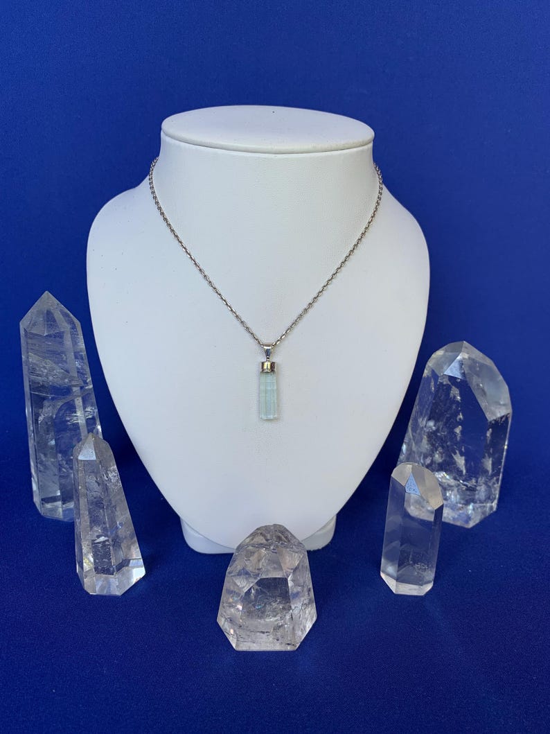 May include: A pale blue, cylindrical crystal pendant hangs from a thin, silver-toned metal chain. The pendant is capped with a small silver-toned metal bail. The necklace is displayed on a white, neck-shaped stand against a solid blue background.