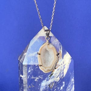 Peut inclure: Un collier en chaîne argent avec un pendentif en forme de tranche d'agate brun clair avec un centre de cristal blanc. Le pendentif est présenté sur un cristal de quartz clair.