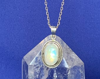 May include: A silver necklace featuring an oval moonstone pendant with a rope-textured border. The pendant hangs from a delicate silver chain, resting against a clear, faceted quartz crystal point that displays internal fractures and light reflections.