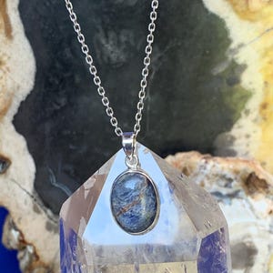 May include: A silver necklace with an oval pendant featuring a blue and gray stone. The pendant is set in a silver bezel and hangs from a delicate silver chain. The necklace is displayed on a clear crystal.