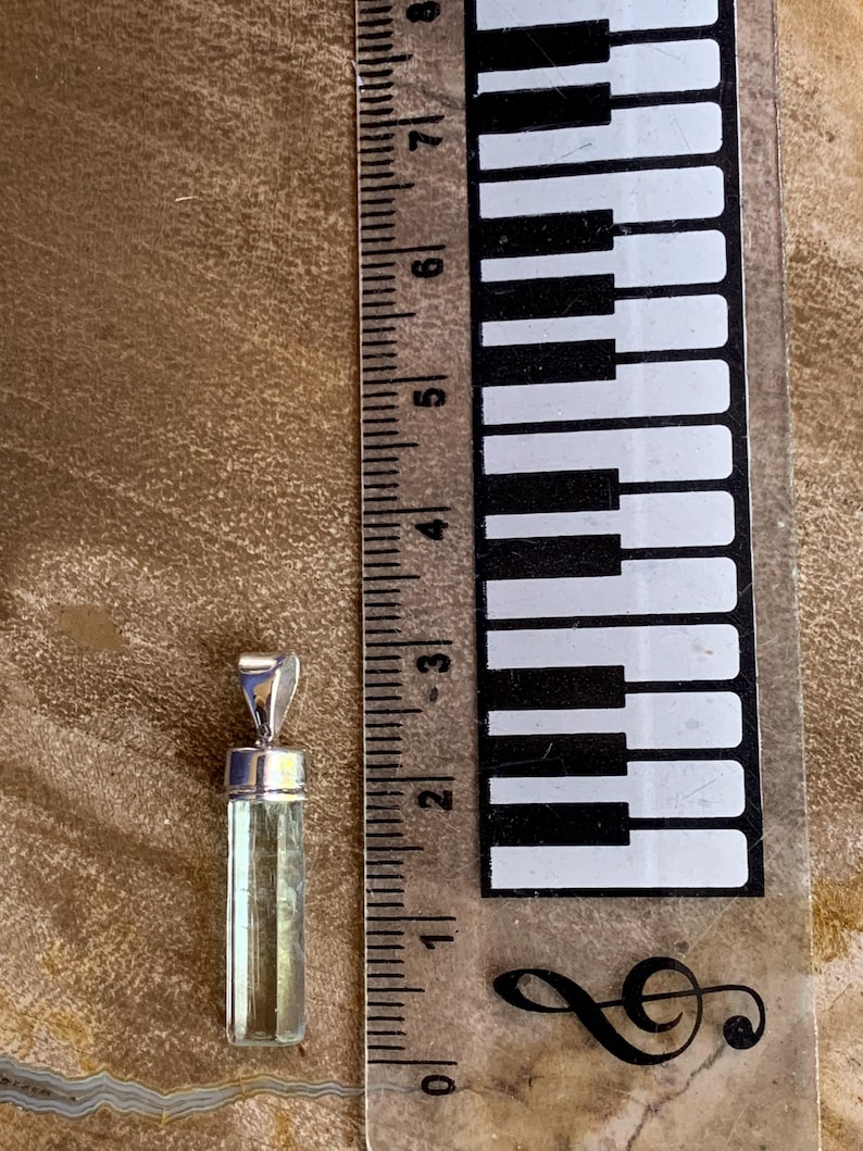 May include: A pale green, rectangular, faceted gemstone pendant with a silver-toned metal bail. The pendant is positioned next to a ruler, showing a length of approximately 1.25 inches from the top of the bail to the bottom of the stone.