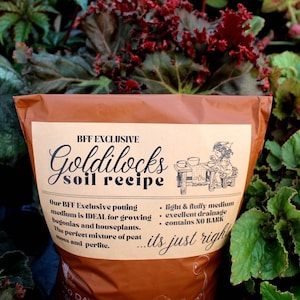May include: A brown bag of potting soil labeled "BFF Exclusive Goldilocks Soil Recipe". The bag features a cartoon illustration of a girl with a watering can and a wheelbarrow. The text on the bag reads "Our BFF Exclusive potting medium is IDEAL for growing begonias and houseplants. The perfect mixture of peat moss and perlite... its just right."
