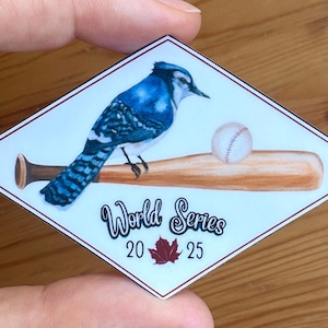 Può includere: Un adesivo a forma di diamante con una ghiandaia azzurra appollaiata su una mazza da baseball e una palla da baseball. Il testo recita "World Series" con i numeri "20 25" e una foglia d'acero rossa.