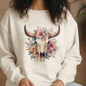 May include: A white sweatshirt with a floral design featuring a bull skull. The skull is surrounded by pink, yellow, and orange flowers.