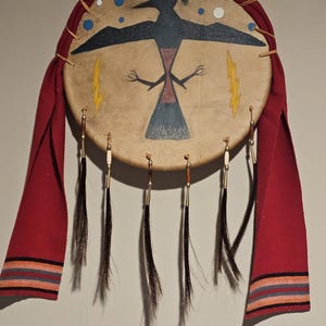May include: A round, brown leather Native American drum with a painted design of a black bird with yellow lightning bolts and blue and white circles. The drum has red tassels and a red fabric border.