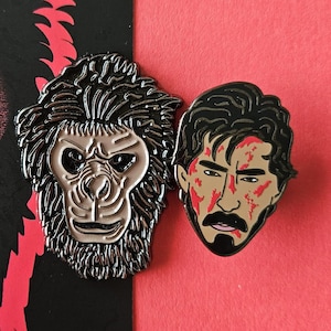 Może przedstawiać: Dwie emaliowane przypinki przedstawiające czarno-białego małpę i mężczyznę z zakrwawioną twarzą. Przypinka z małpą ma czarne tło, a przypinka z mężczyzną ma czerwone tło.