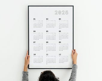 May include: A black framed 2025 calendar with a white background. The calendar displays the months from January to December, with each month's days arranged in a grid. The calendar is held by a person.