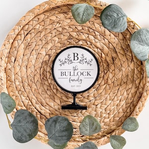 May include: A round, black and white family sign with the text "the BULLOCK family" and "EST. 2022". The sign is on a black stand, set on a woven, circular placemat, and surrounded by green eucalyptus leaves.