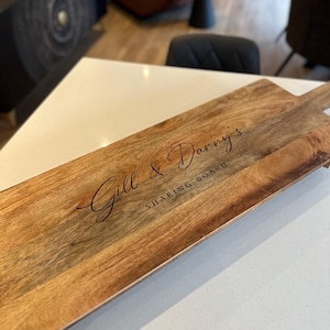 May include: A wooden sharing board with a handle, displaying the text "Gill & Danny's SHARING BOARD". The board has a natural wood grain and is rectangular. It rests on a white surface, showcasing its warm tones and design.