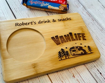 Puede incluir: Una bandeja rectangular de bambú para aperitivos con un hueco circular para una bebida, grabada con láser con el texto "Robert's drink & snacks" y "VanLIFE" sobre un gráfico de una autocaravana rodeada de pinos y pequeñas estrellas.