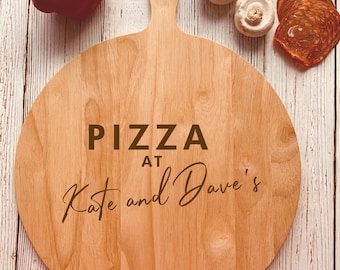 May include: A round wooden pizza serving board with a handle. The board is engraved with the text "Pizza at Kate and Dave's".