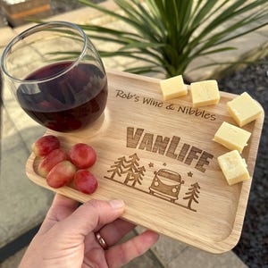 May include: A wooden serving tray with a glass of red wine, grapes, and cheese cubes. The tray is engraved with "Rob's Wine & Nibbles" and "Vanlife" with a camper van graphic. The tray is held by a person.